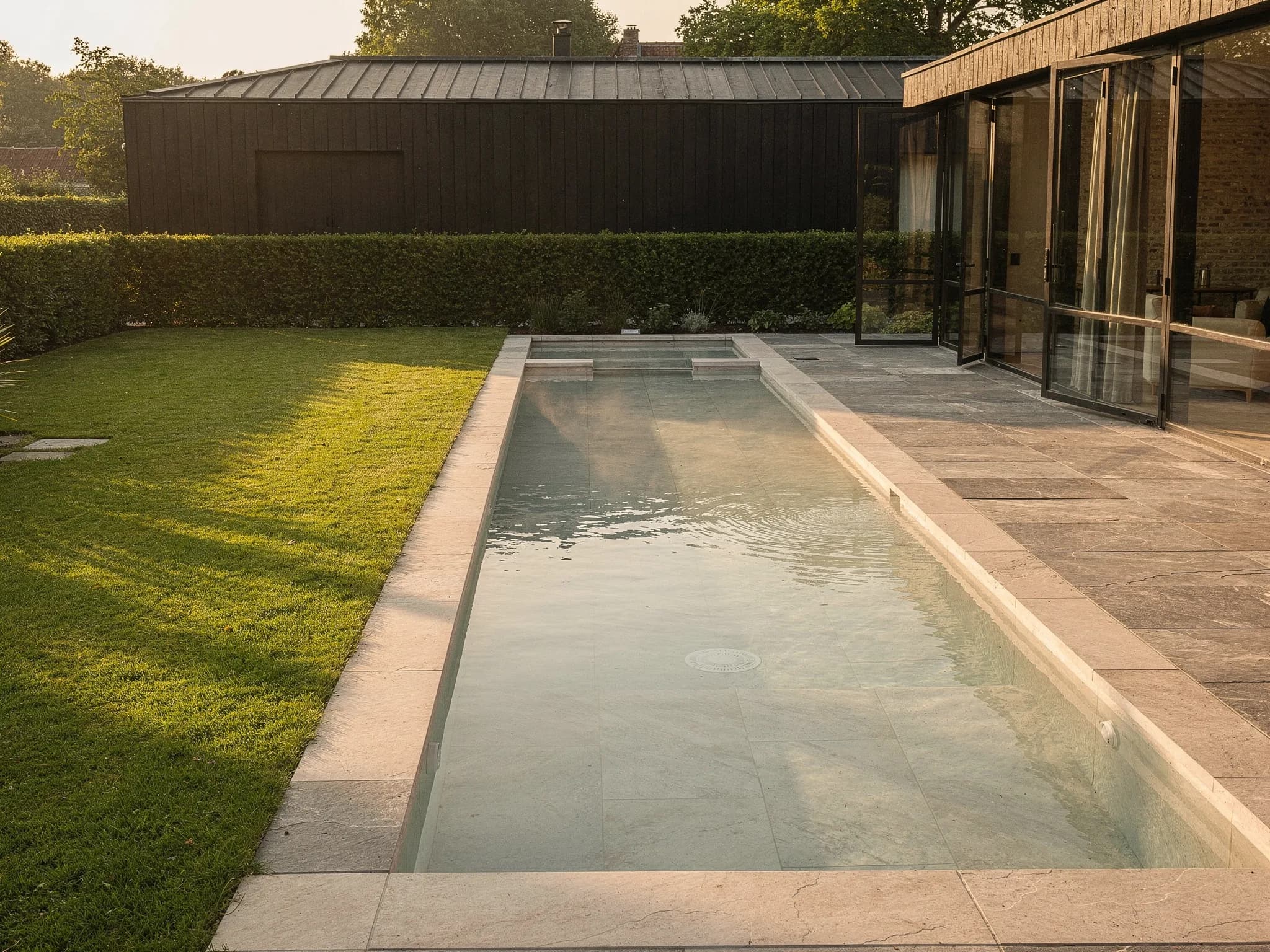 Conceptbeeld van een langgerekt baanbad met lichte keramische tegels naast een zwart houten schuurwoning en gazon