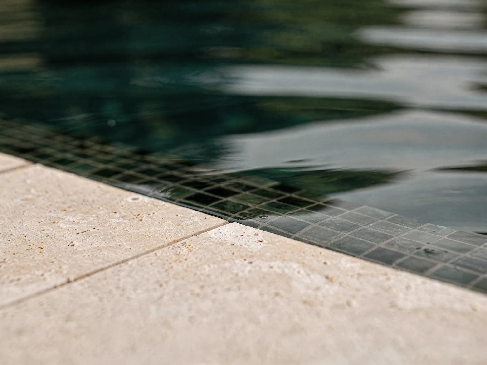 Detail van kalm zwembadwater tegen een kalkstenen boordsteen met donkere mozaïekwaterlijn en bronzen accent